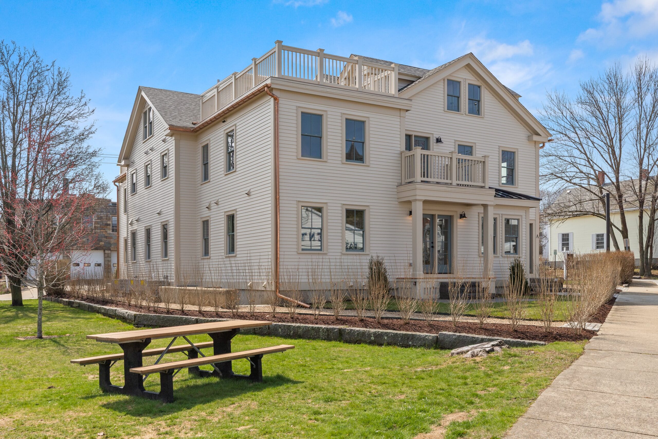 A three-story beige house at One Jewett Street features a small balcony and rooftop deck, surrounded by a yard with a picnic table on the lawn, all under a blue sky.