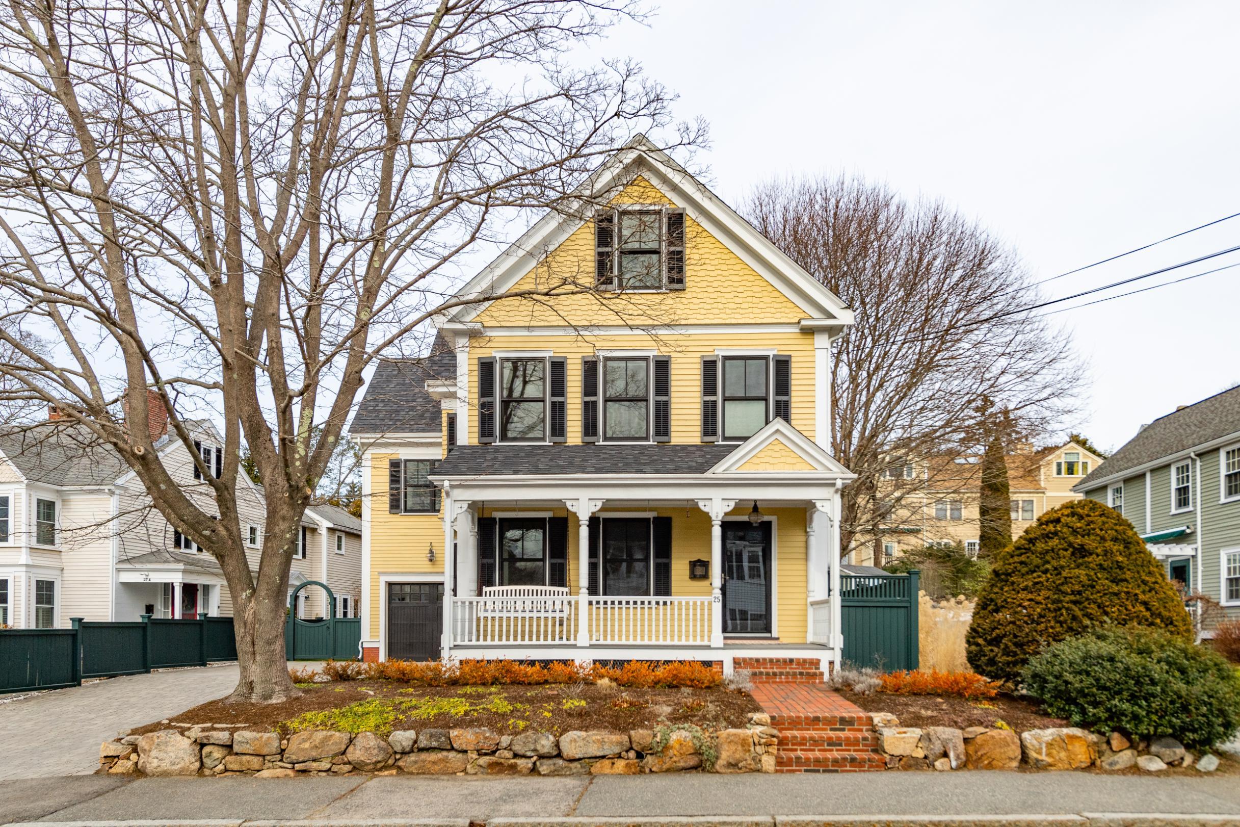 A two-story yellow house with white trim, front porch, and black shutters, surrounded by leafless trees and shrubs, sits behind a stone-lined garden and sidewalk.