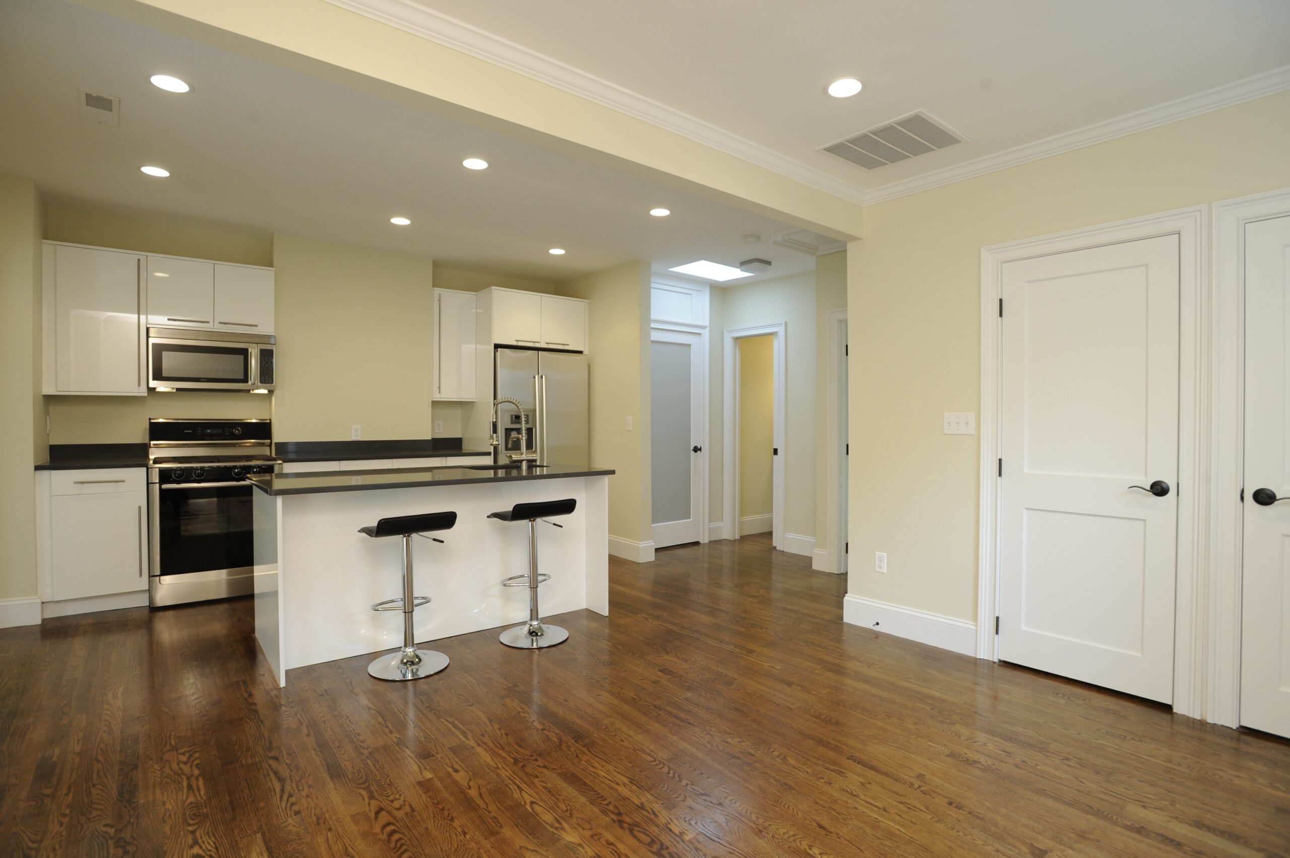 Modern kitchen on Greenview Avenue with stainless steel appliances, white cabinets, black countertops, two bar stools, and hardwood floors, adjacent to a hallway and two closed white doors.