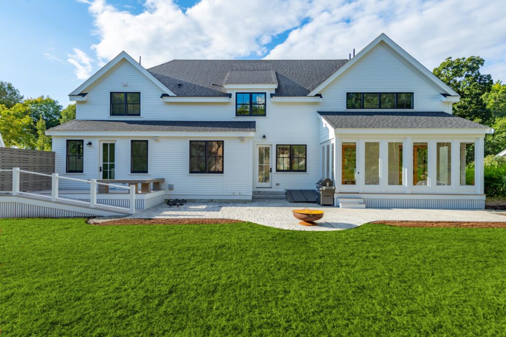 A modern two-story white house with large windows, a spacious patio, a fire pit, and a well-manicured lawn under a partly cloudy sky.